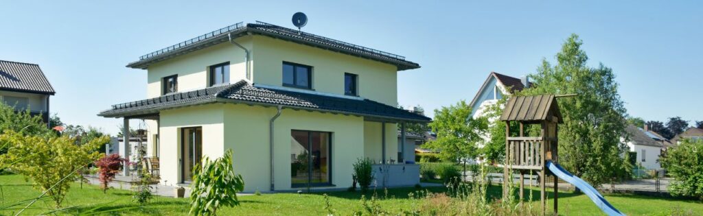 Auf dem Bild ist ein neugebautes Walmdachhaus zu sehen im Stadtvillen-Stil. Die Fassade ist in weichem Zitronengelb.